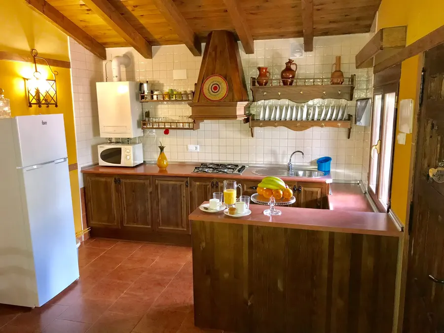 Cocina rústica de una cabaña con muebles de madera, campana de obra, vitrina de platos y barra abierta al salón