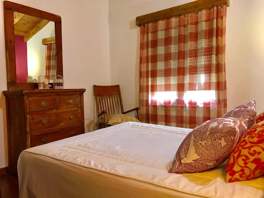 Dormitorio de una cabaña con cómoda de madera antigua con espejo, silla de enea y cortinas de cuadros rojos