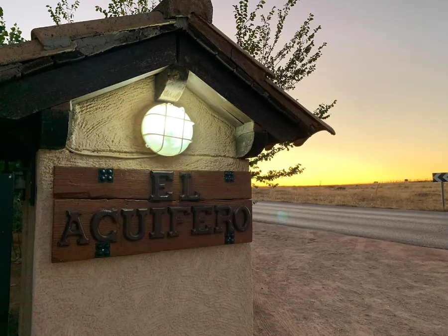 Cartel de hierro de Cabañas El Acuífero sobre el pilar de piedra de la entrada, con el farol encendido y la puesta de sol sobre el llano