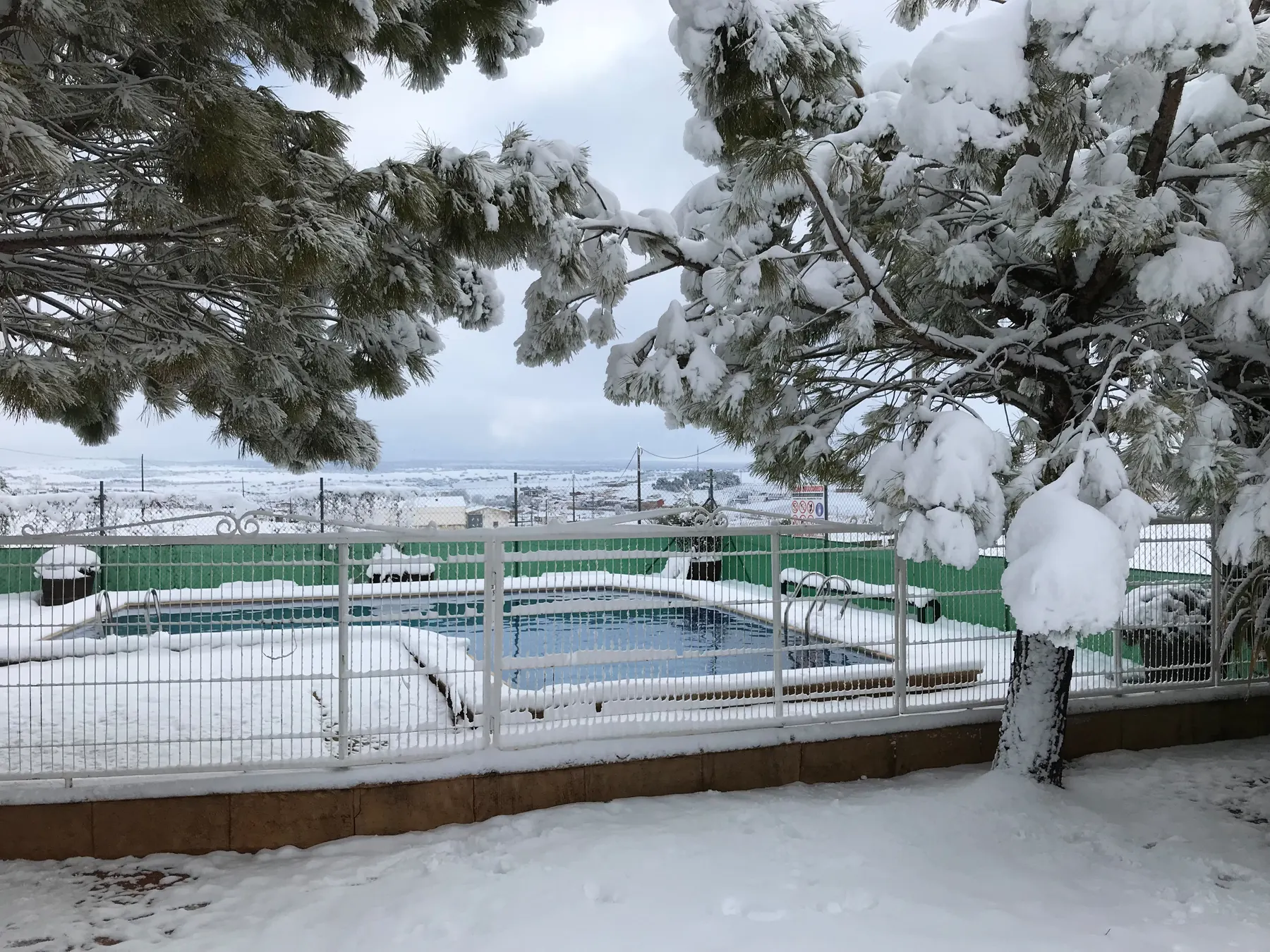 La piscina de El Acuífero nevada en invierno, entre pinos cargados de nieve y con el campo blanco al fondo