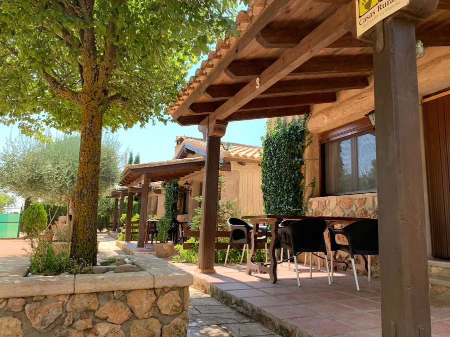 Fila de porches de madera de las cabañas de El Acuífero, con mesas y sillas bajo las vigas y un muro de piedra en primer plano