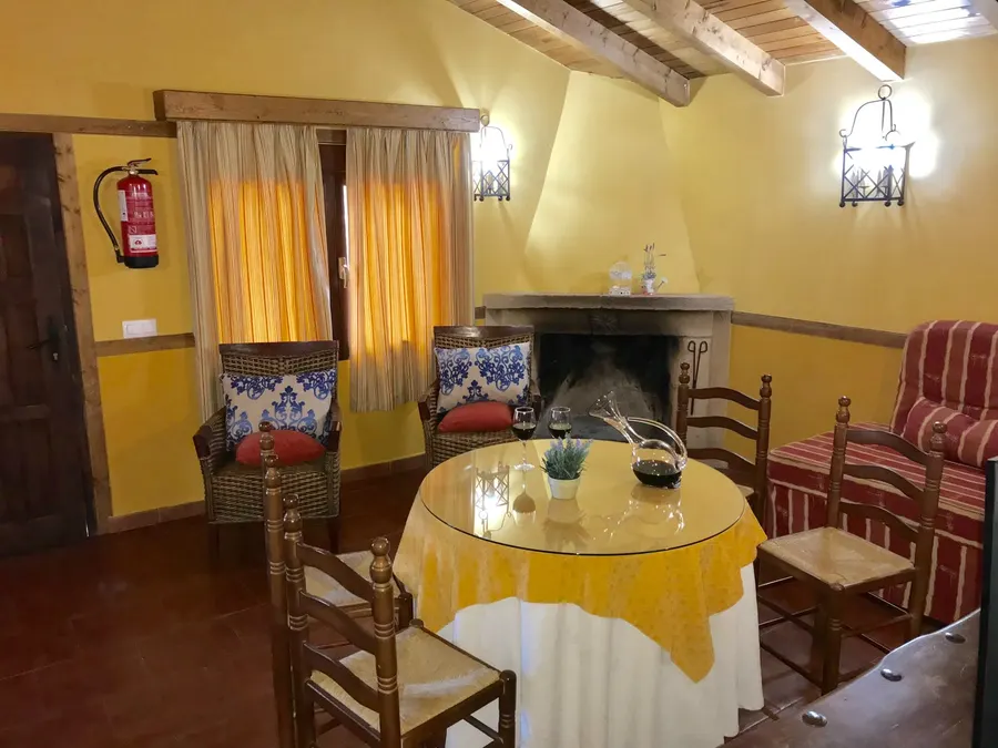 Salón de una cabaña de El Acuífero con chimenea de leña, vigas de madera vistas, mesa camilla y butacas de mimbre