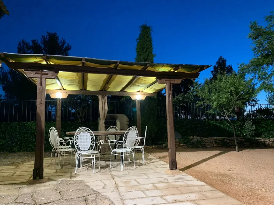 Pérgola de madera iluminada de noche en la zona común de El Acuífero, con mesa larga, sillas de forja y la barbacoa al fondo